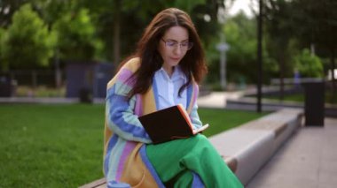 Uzun saçlı, gözlüklü genç bir kadın iş programını planlıyor. Fikirlerini not defterine bir kalemle yazıyor. Parktaki bir bankta oturuyor..