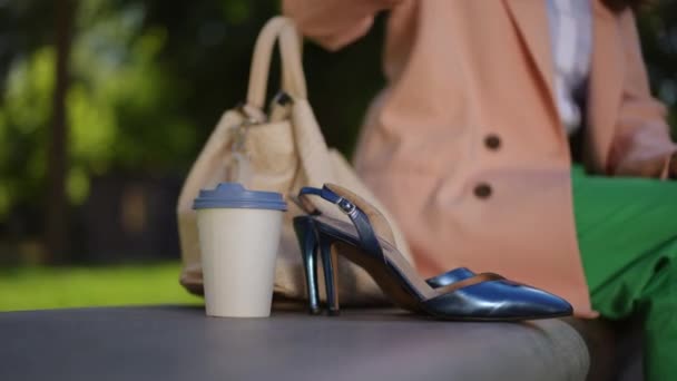 ある女性が公園のベンチに座り 青い閉じたサンダルの隣にレザーバッグを置く 女性は彼女のサンダルを手に取ってそれらを置きます クローズアップ ボディパーツ — ストック動画
