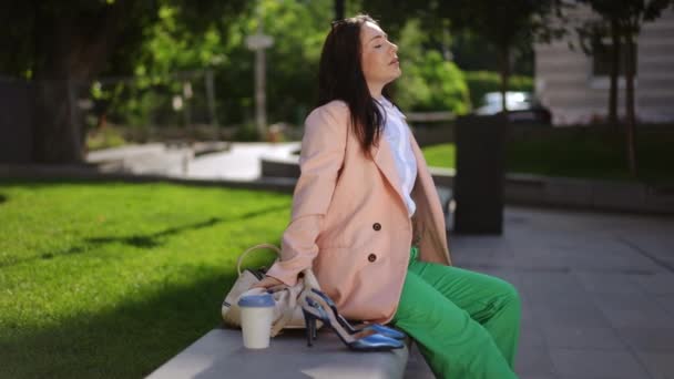 在一个阳光灿烂的日子里 一位时尚的女商人坐在公园的长椅上休息 旁边的长椅上有一个皮包和一双工作鞋 — 图库视频影像