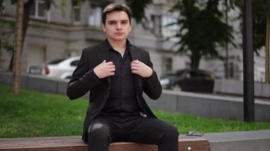 Takım elbiseli genç bir adam bir şehir parkında bankta otururken ceketini düzeltir ve gülümser. Adam kameraya bakar ve gülümser.