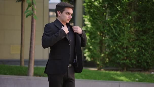 黒い正式なスーツを着た若者はジャケットを調整してボタンをかけ 街の木々の背景に立っている — ストック動画