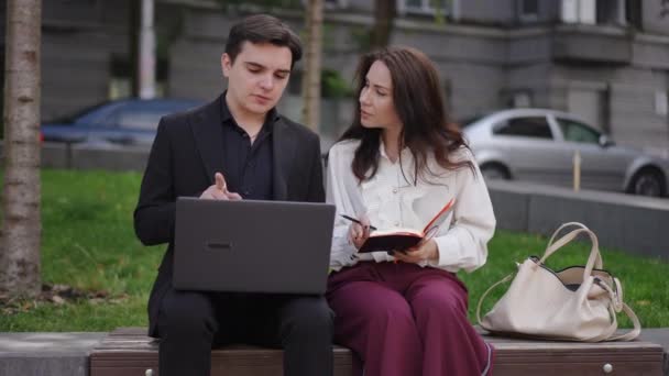 男性は ソフトウェアを正しく使用する方法をノートパソコンに女性を示しています 女性は注意深くリストアップし メモ帳に書き込みます カップルは市街地のベンチに座っている — ストック動画