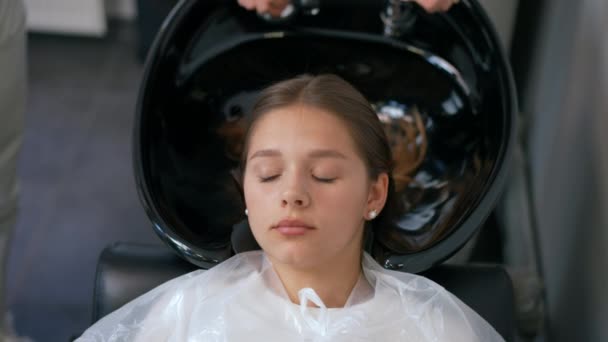 女の子はシンクでヘアメイドの椅子に横たわっています 彼女の後ろには 美容師が洗面台に水を流し 女の子の髪を滑らかにします クローズアップ — ストック動画