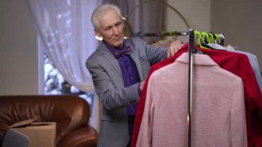 Yaşlı bir erkek moda tasarımcısı, modern bir evin odasındaki bir elbise dolabının askılarında asılı kıyafetlerini inceliyor.