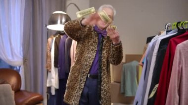 Şık bir leopar desenli ceketli abartılı yaşlı bir moda tasarımcısı modern bir evin soyunma odasında duruyor, gülümsüyor, para saçıyor ve dans ediyor..