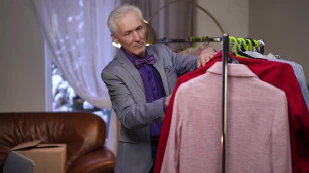 高齢の男性ファッションデザイナーは 現代の家の部屋でモバイルワードローブハンガーのハンガーに掛かって彼の服を調べます — ストック動画