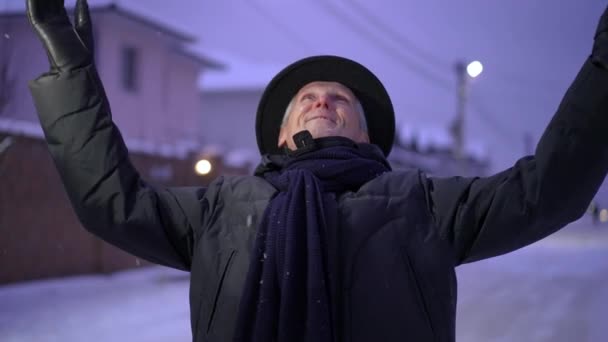 明るい帽子を被った年配の男性が手を挙げて笑顔を浮かべ 冬の雪だるまに立っている間に空を見上げます 夕方になると雪が降る 男は手を下げ 微笑み 見ている — ストック動画