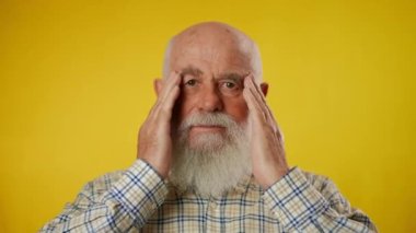 Tişörtü olan, başı ağrıyan ve sarı arka planda izole edilmiş elleriyle şakaklarını ovuşturan yaşlı, gri sakallı bir adamın portresi. Bir adam kameraya bakıyor. Yakın plan..