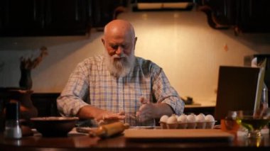 Tişörtünde sakalı olan kel kafalı yaşlı bir adam mutfak masasında oturuyor ve elleriyle hamuru yuvarlıyor. Bir adam masanın üstünde duran bir kitaba bakar.