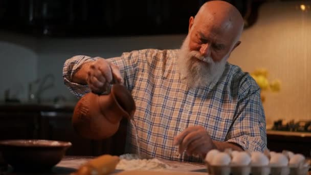 ひげ付きの老人が台所のテーブルに座っている間に生地をひざまずく 男は粘土のジャグから水を小麦粉に汲み 手でそれをひざまずく クローズアップ — ストック動画