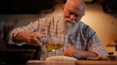 Yakın plan. Gömleğinde sakalı olan yaşlı kel bir adam saplı cam bir konteynırdan bitkisel yağ dökerek dikdörtgen bir hamur parçasının üzerine döküyor ve eliyle ovuyor. Hamur bir tahtanın üzerinde yatıyor.