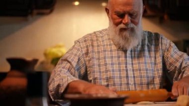 Yakın plan. Yaşlı, sakallı, gömlekli bir adam tahta bir tahtanın üzerinde merdaneyle hamuru yuvarlıyor. Bir adam hamura un serpiyor. Ev mutfağında hamur tatlısı yapıyorum.