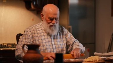Ev mutfağında hamur tatlısı hazırlama süreci. Yaşlı, sakallı, gömlekli bir adam tahta bir tahtanın üzerinde merdaneyle hamuru yuvarlıyor. Bir adam hamurun üzerine un serper.