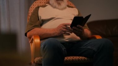 Tişört ve kot pantolon giymiş yaşlı, tanınmayan sakallı bir adam elinde tuttuğu cep telefonuyla internetten içeriği izlerken gülüyor, rahat bir şekilde sallanan bir sandalyede oturuyor.