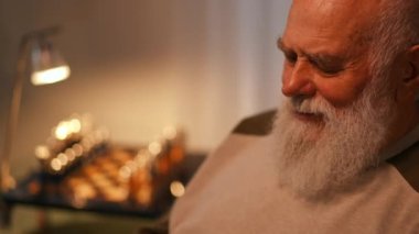 Yakın plan. Tişörtlü yaşlı sakallı bir adam oturma odasında sallanan sandalyede otururken elinde tuttuğu alete bakarken gülüyor.