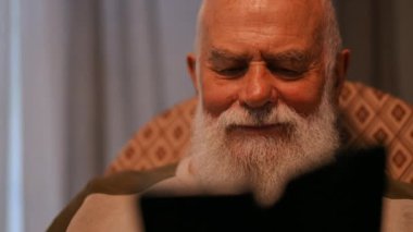 Yakın plan. Tişörtlü yaşlı sakallı bir adam evinin rahat oturma odasında sallanan sandalyede otururken elinde tuttuğu cep telefonuna bakarken gülüyor.
