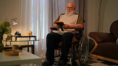 Yalnız, engelli bir adam tişört giyiyor, elinde tuttuğu çerçeveli fotoğrafa bakıyor, evinin rahat oturma odasında tekerlekli sandalyede oturuyor.