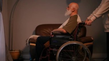 Tekerlekli sandalyede oturan yaşlı engelli bir adam, tanınmayan bir kişi tarafından perdeli pencereye doğru sürülür.