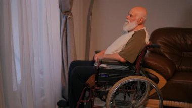Yalnız, sakallı, sakallı, tişörtlü yaşlı bir adam oturur ve perdeli pencereden bakar ve elini sakalının üzerinden uzatır. Tekerlekli sandalyede, evinin rahat oturma odasında.