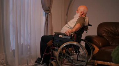 Tekerlekli sandalyede oturan yaşlı, sakallı, sakallı, sakallı, sakallı biri. Engelli adam perdeli pencereden dışarı bakar ve kollarını sallayarak onları gerer.