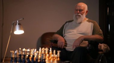 Tekerlekli sandalyede oturan yaşlı sakallı bir adam satranç oynarken taktik ve strateji düşünür.