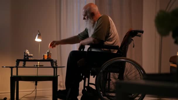 在一个灯光昏暗的客厅里 一个孤独的 留着胡子的残疾老人坐在轮椅上下棋 — 图库视频影像