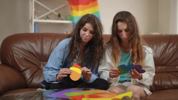 リビングルームのソファーに座っている2人の少女は それぞれ3枚のマルチカラーのボール紙のハートを拾い カメラに持って行きます 女の子は微笑み 虹の背景からカメラを見る — ストック動画