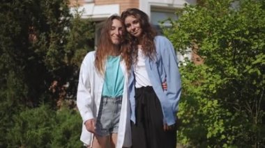 İki kız, güneşli bir yaz gününde yeşil ağaçların arasında bir şehir parkında birbirlerine sarılıyorlar. Kızlar gülümsüyor ve kameraya bakıyor.