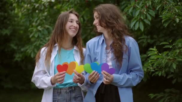 两个女孩手里拿着彩色纸板的心 站在树旁的室外 女孩子们在鬼鬼祟祟地笑着 看着摄像机 — 图库视频影像