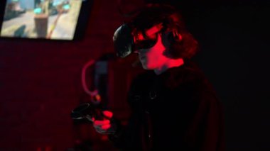 Yakın plan. Kırmızı ışıklandırmalı bir VR kulübünün karanlık bir odasında, VR kasklı bir genç elinde kontrolörlerle bir video oyunu oynuyor.