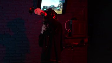 VR gözlük takan ve elinde kontroller tutan genç bir adam video oyunu oynarken okçuluğu taklit ediyor. Kırmızı ışıklandırmalı karanlık bir odada, duvara asılı bir monitör var.