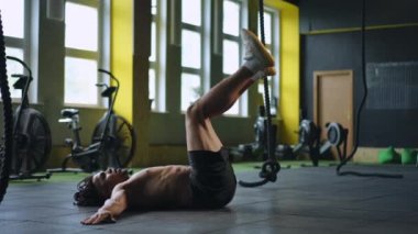 Melez bir ırk modern bir spor salonunda sırt üstü yatarken bacaklarını kaldırır. Adam kollarını yana açmış sırt üstü yatıyor.