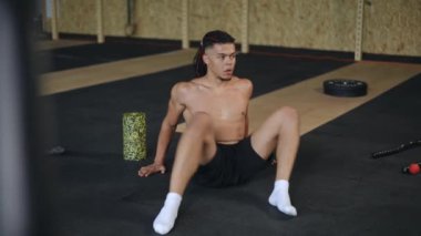 Kafasında rasta olan melez bir adam spor salonunda yerde otururken dinamik bir kalça esneme egzersizi yapıyor.