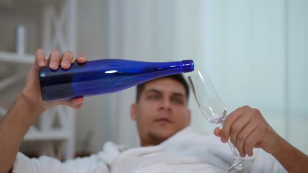 スローモーション クローズアップ 白いバスローブの若者が青い狭いボトルからワイングラスに白ワインを飲み込み 寝室のベッドに横たわっている — ストック動画