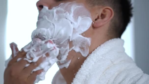 クローズアップ 白いバスローブの男は寛大に浴室に立っている間彼の顔に泡を剃ることを適用します — ストック動画