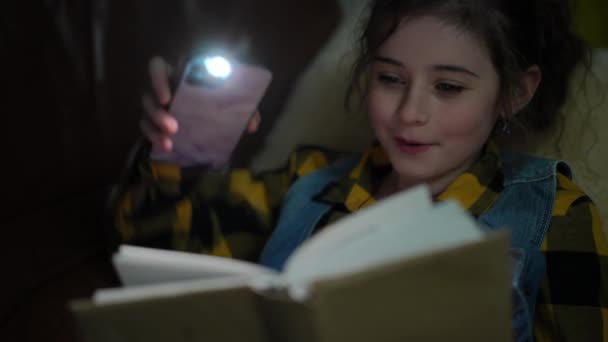 Zbliżenie Szczęśliwa Dziewczyna Czytająca Książkę Pomocą Latarki Telefonu Komórkowego Leżąca — Wideo stockowe