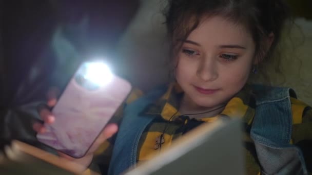 クローズアップ 少女は興味深い本を検討しています 本のページを照らす 彼女の携帯電話からの懐中電灯で 現代の家のリビングルームのソファーの暗闇に横たわる — ストック動画