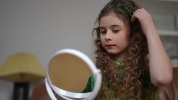 緑色のドレスの美しい小さな女性は 彼女の髪のクリップと髪をまっすぐにまっすぐにして 小さな鏡で自分自身を見ています モダンなリビングルームでソファーに座っている少女 — ストック動画