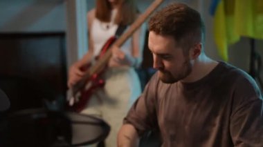 Yakın plan. Bir adam oturur ve ustaca elektronik bateri çalar ve tanınmayan bir kız bas gitar çalar. Bir müzik okulundaki ritm bölümünün provaları bayrağın arka planında