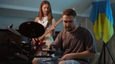 Ağır çekim. Bir adam elektronik davul setini çalmaya başlar ve genç bir kız bas gitar çalmaya başlar. Bir müzik okulundaki ritm bölümünün provaları bayrağın arka planında
