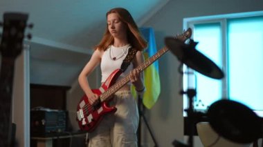 Genç bir kız, bir müzik okulunun binasında Ukrayna bayrağının arka planında dikilirken enerjik bir şekilde bas gitar çalar.