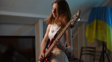 Genç kızıl saçlı bir kız, duvarında Ukrayna bayrağı olan bir müzik okulunda dikilirken enerjik bir şekilde bas gitar çalıyor.