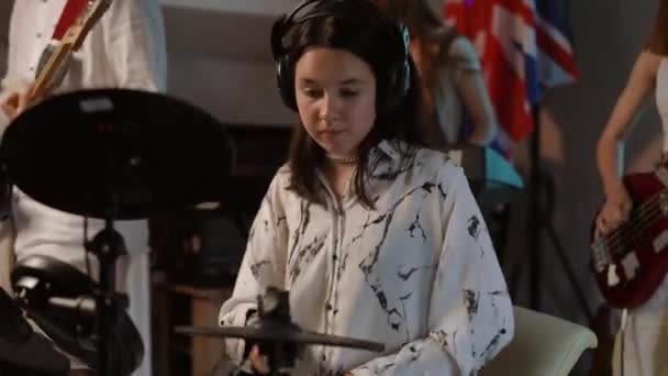 在音乐学校的教室里排练的一个音乐小组 在前景中 一个戴耳机的女孩会玩一个电子鼓包 在背景下 难以辨认的青少年在玩耍 — 图库视频影像