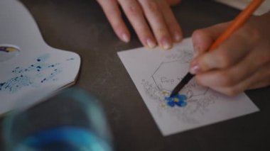 Yakın plan. Bir kadın bir apartmanın oturma odasındaki masada otururken boya fırçası ve suluboya ile bir tebrik kartı çiziyor. DIY Anneler Günü tebrik kartı