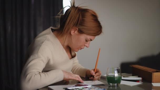 一位红头发的女士坐在客厅的桌子旁 她用画笔仔细地画了一张带有水彩画的贺卡 Diy贺卡 — 图库视频影像
