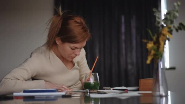 一个女人用画笔画水彩画 一个女人坐在客厅的桌子旁 周末和假日的家庭业余爱好 — 图库视频影像