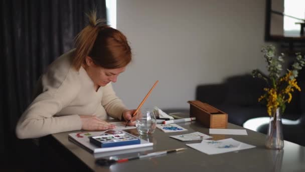 慢动作一个女人坐在客厅的桌子旁 微笑着用水彩画贺卡 Diy贺卡作为度假和重要约会的嗜好 — 图库视频影像