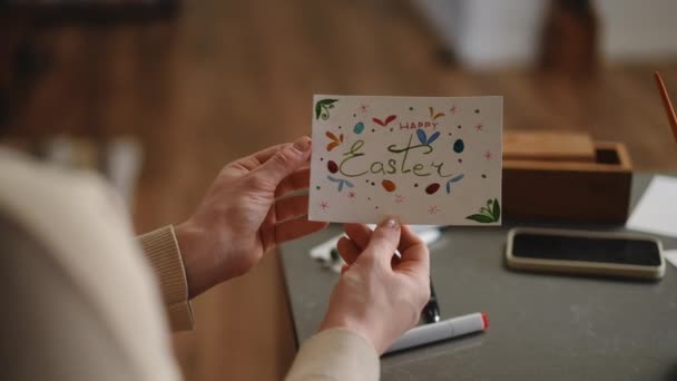 慢动作一个女人拿着一张复活节贺卡 她坐在客厅的桌子旁 上面画着彩色记号和水彩画 Diy复活节卡片 背对背射击 — 图库视频影像