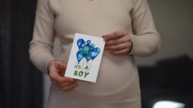 Ağır çekim. Elinde doğmamış çocuğu için bir doğum günü kartı tutan tanınmamış hamile bir kadın. Kapalı alanda dururken renkli kalemler ve suluboya kalemlerle çizmiş. DIY tebrik kartı
