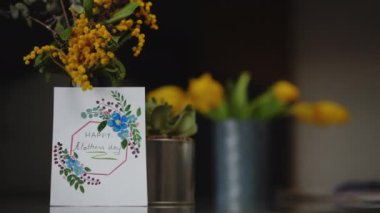 Yakın plan. İçinde Anneler Günü kartı olan bir buket bahar çiçeği kapalı bir masada duruyor.
